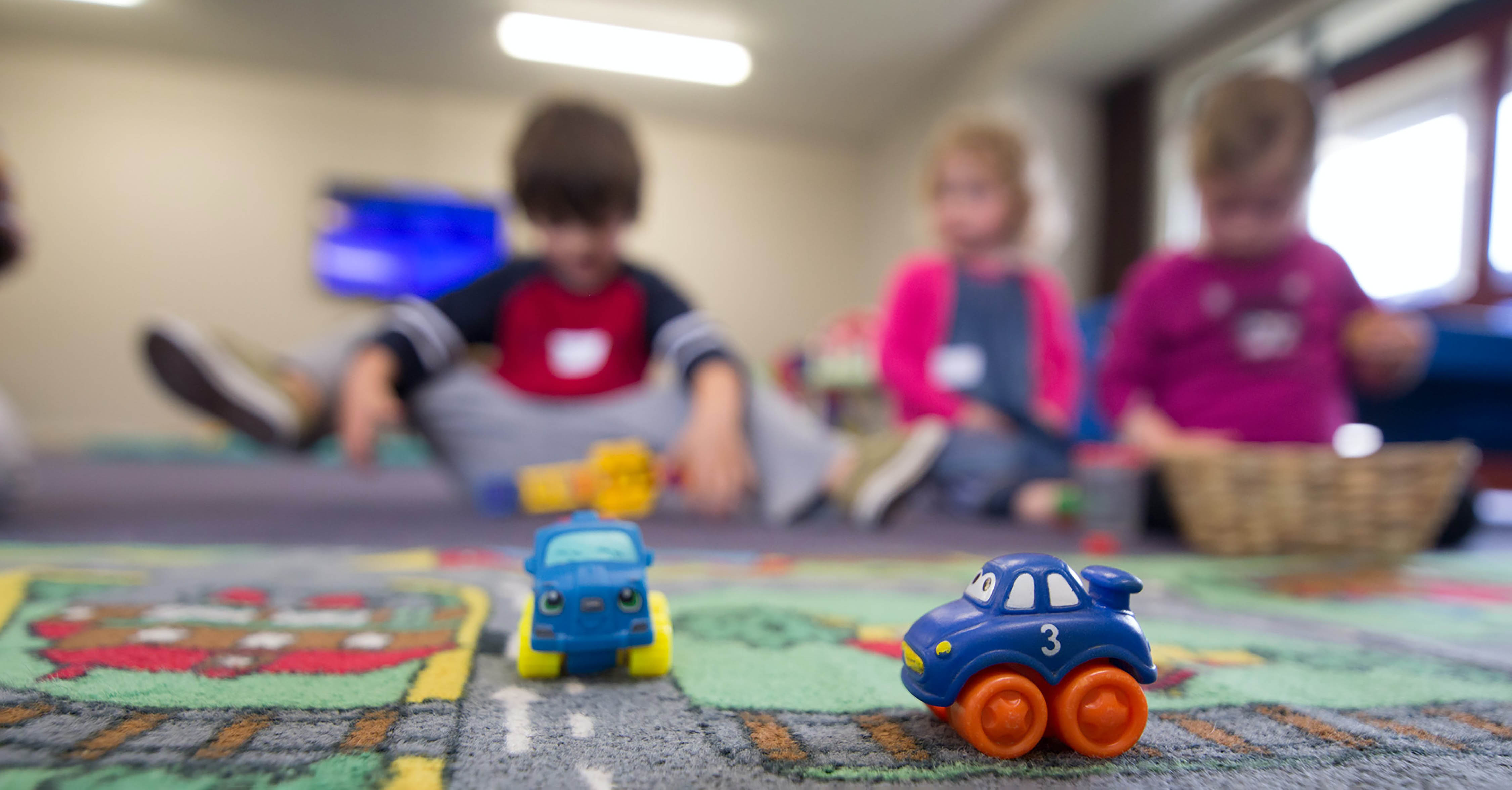 Three young children play on a carpet with toy cars in a classroom.
